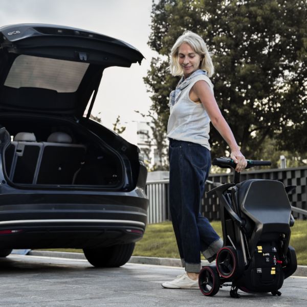 A woman stands outdoors beside an open car trunk, pushing a folded Enhance Mobility iMOVE AirCarbon. Trees, grass, and a fence are visible in the background.