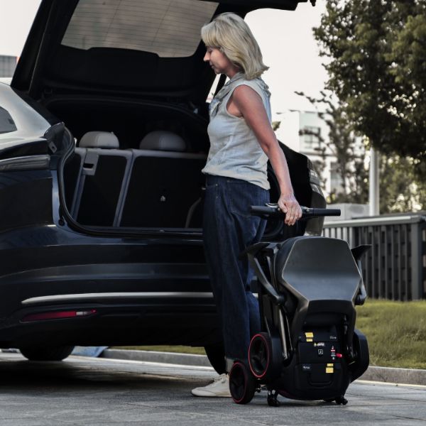 A woman stands beside an open black car trunk in a parking lot, holding a compact Enhance Mobility iMOVE AirCarbon. The lift is black with small red wheel accents and a handlebar, shown in use outdoors.