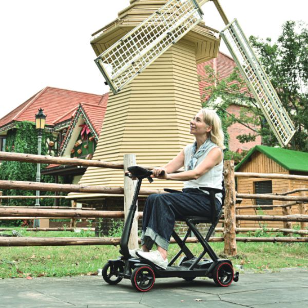 A woman riding a Enhance Mobility iMOVE AirCarbon with red-rimmed wheels in front of a large windmill structure outdoors.