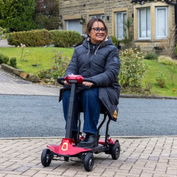 A woman wearing a dark puffer jacket and jeans sits on the Genie Lightweight Folding Mobility Scooter on a paved driveway in front of a residential house.