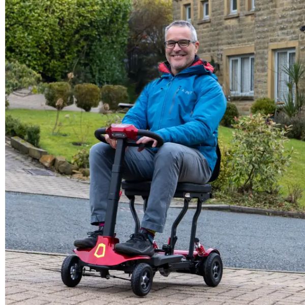 A man wearing a blue jacket and grey trousers sits in Genie Lightweight Folding Mobility Scooter on a paved driveway in front of a stone house and green garden.