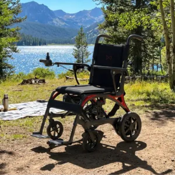 Monarch Mobility Ezi Fold Carbon Ultra-Lightweight Folding Electric Wheelchair, parked outdoors on a dirt path near a lake with mountains and trees in the background.