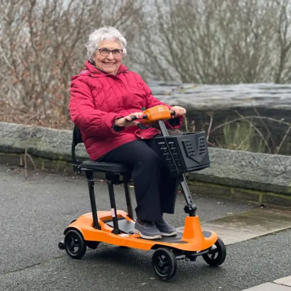 An older woman wearing a bright pink jacket and black trousers sits on an orange Monarch Mobility Genie Ultralight Ultra-Portable Folding Scooter on a paved path with a stone wall and bare trees in the background.
