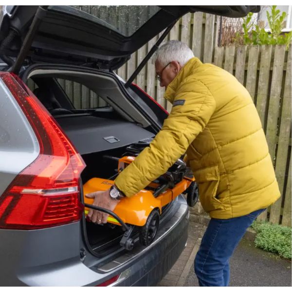 A man in a yellow puffer jacket loads an orange Monarch Mobility Genie Ultralight Ultra-Portable Folding Scooter into the open trunk of a silver car, with a wooden fence in the background.
