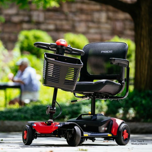 A red Pride Go-Go Elite Traveller Gen 3 4-Wheel Scooter, parked outdoors on a paved path with a blurred garden and stone wall background.