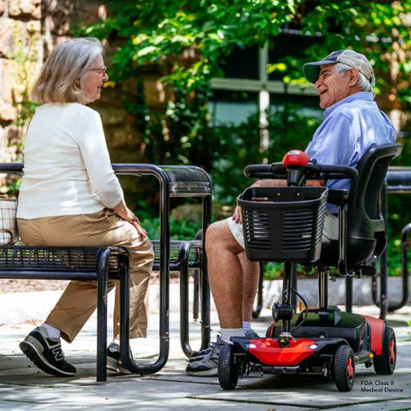 An older man sits in a red Pride Go-Go Elite Traveller Gen 3 4-Wheel Scooter on an outdoor patio, talking to an older woman seated on a bench nearby. Lush green foliage and stone walls are visible in the background.