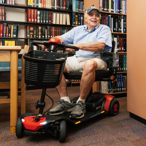 An older man wearing a light blue polo shirt and khaki shorts sits in a red and Pride Go-Go Elite Traveller Gen 3 4-Wheel Scooter inside a library with bookshelves in the background.