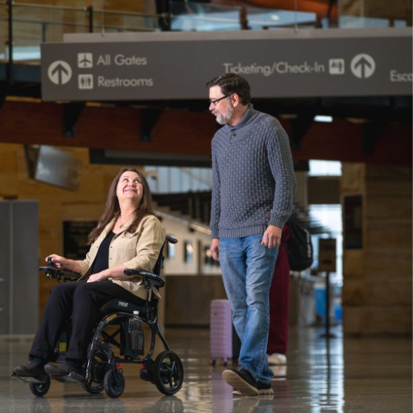 Person using the Pride Jazzy Carbon 27X Lightweight Folding Wheelchair and another person walking in an airport terminal.