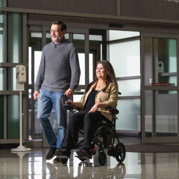 Man pushing a woman in the Pride Jazzy Carbon 27X Lightweight Folding Wheelchair through a building entrance.