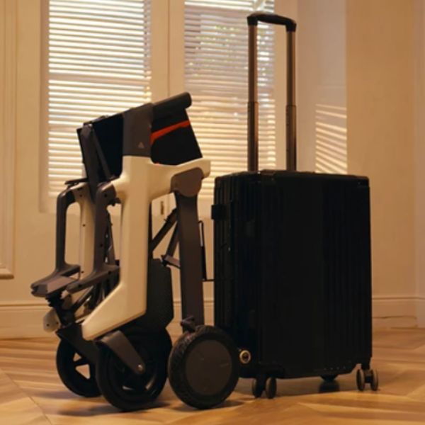 Roamate 3-in-1 Rollator Transport Electric Wheelchair is shown next to a black hard-shell rolling suitcase with an extended telescoping handle in an indoor room with window blinds and wood flooring.