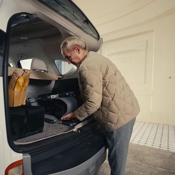 An older man in a beige quilted puffer jacket and gray pants is loading a folded Roamate 3-in-1 Rollator Transport Electric Wheelchair