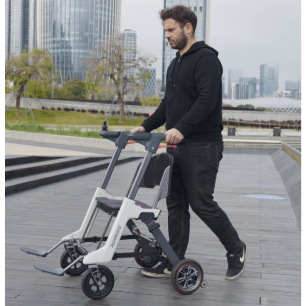 A man in a black hoodie and jeans pushes a Roamate 3-in-1 Rollator Transport Electric Wheelchair Black across a paved outdoor walkway, with a city skyline in the background.