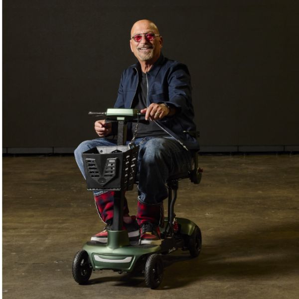 A man wearing red-tinted sunglasses sits on the Monarch Mobility Smarti Plus auto-folding mobility scooter photographed in a dimly lit studio setting.