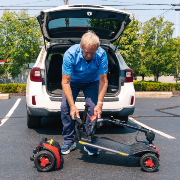 A man in a blue polo shirt and dark pants is shown outdoors in a parking lot, maneuvering a Pride Go-Go Elite Traveller® Gen 3 Platinum 4-Wheel Mobility Scooter in front of an open white SUV trunk.
