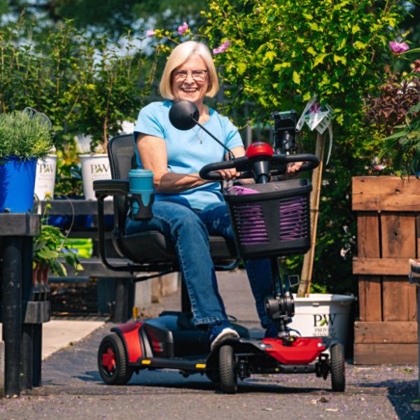 A woman wearing glasses and a light blue shirt sits smiling in a Pride Go-Go Elite Traveller® Gen 3 Platinum 4-Wheel Mobility Scooter outdoors in a garden setting with plants and wooden crates in the background.