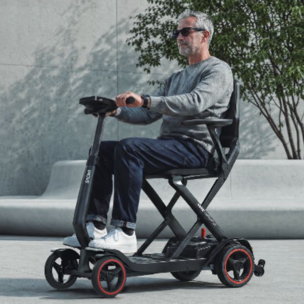A man wearing sunglasses sits on a Enhance Mobility iMOVE AirCarbon with a seat and handlebars in an outdoor urban setting with a concrete wall and tree in the background.